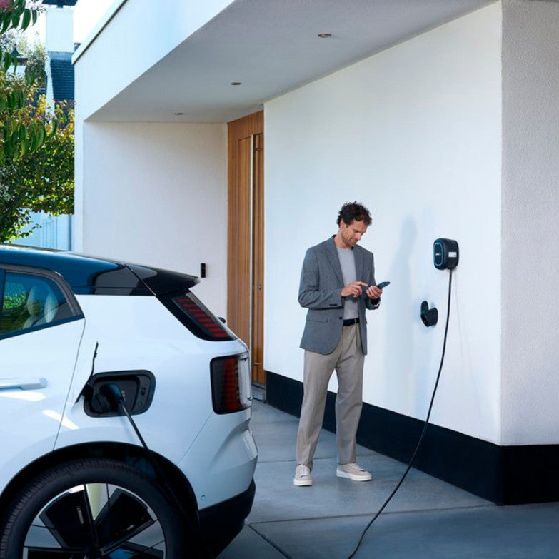 Weißes Elektroauto, das an einer Wallbox neben einem Wohnhaus angeschlossen ist.